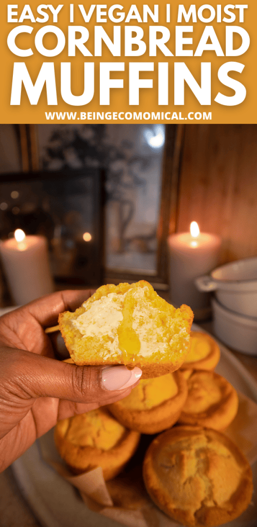 A hand holding a half-eaten vegan cornbread muffin with a moist interior, surrounded by more cornbread muffins on a plate with candles in the background.