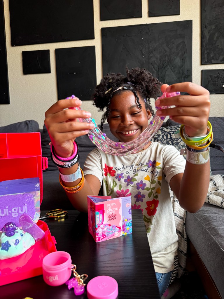A girl smiles while holding up a colorful, beaded bracelet, surrounded by craft supplies and a red storage box.