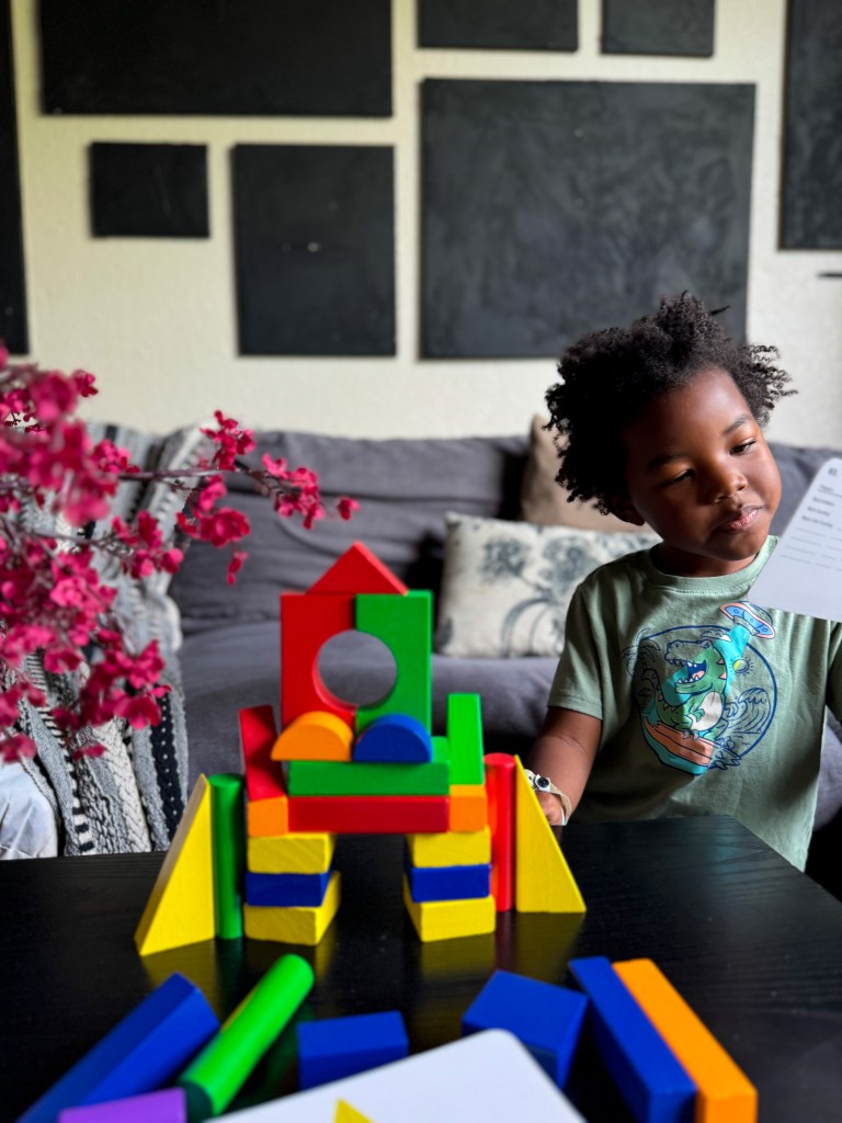 A child playing with colorful building blocks, creating a structure, in a cozy living room setting with plants and artwork in the background.