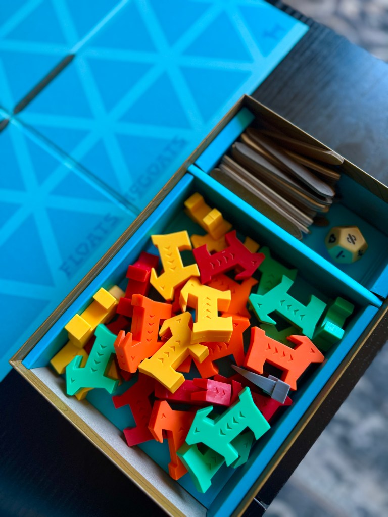 A colorful assortment of game pieces in various shapes, including red, yellow, green, and orange animals, inside a game box alongside wooden sticks and dice. The game board's blue geometric design is visible in the background.