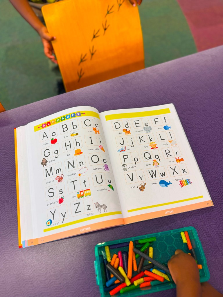 An open book displaying the alphabet with colorful illustrations for each letter, placed on a purple table, alongside a container of colorful crayons and a child's hand reaching for them.