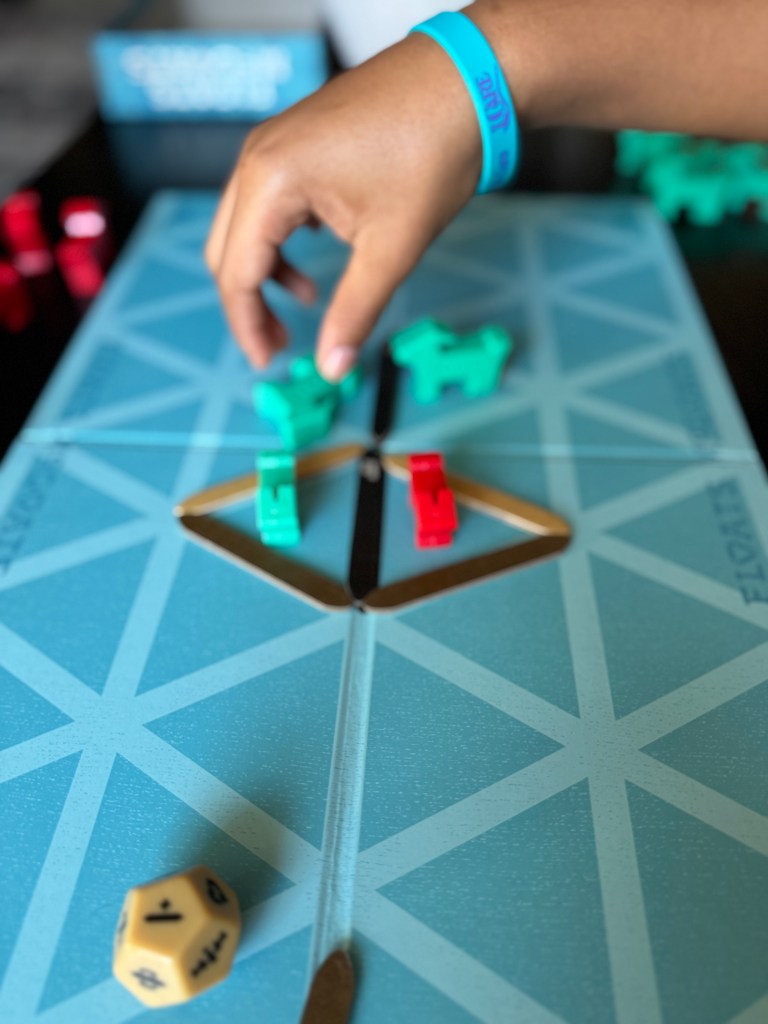 A person's hand reaching for game pieces on a vibrant blue game board, with green and red figures and a yellow die visible.