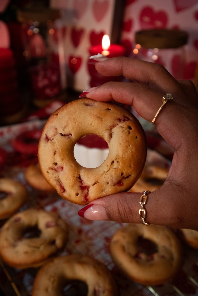 A hand holding a fresh donut with a hole in the center, surrounded by more donuts on a decorative tray, set against a romantic background with candles and heart patterns.