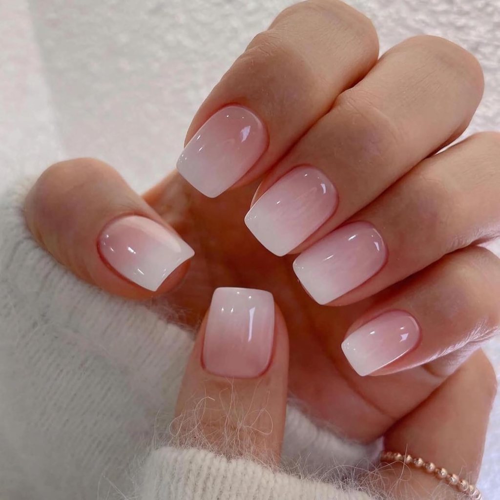 A close-up of a hand with neatly manicured nails featuring a gradient pink and white ombre effect, resting on a soft, white fabric.