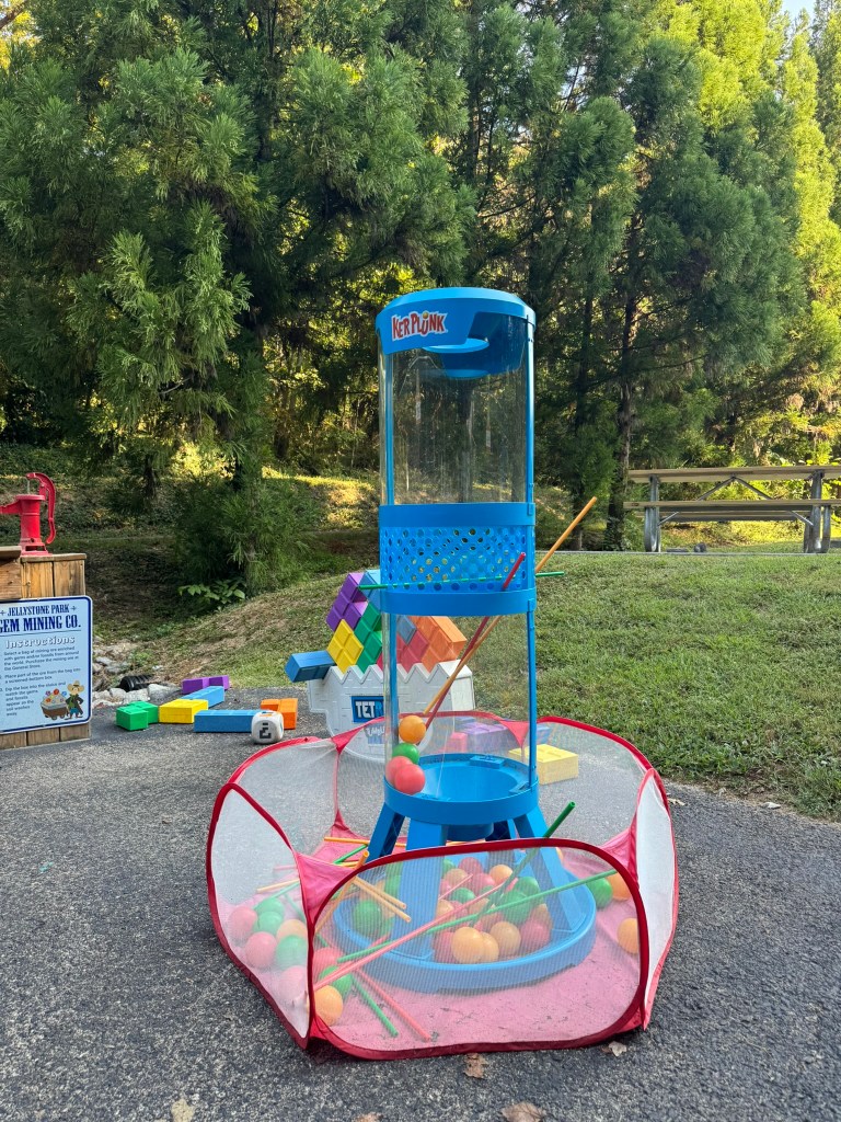 A colorful outdoor game setup featuring a blue tower with a mesh top and a red circular enclosure. The tower has plastic balls inside, and colorful building blocks are scattered nearby on a paved surface, surrounded by trees.