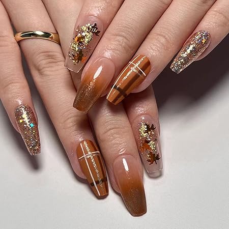 A hand with long, glossy nails featuring a warm autumn color palette of browns and golds. The nails are decorated with glitter, metallic accents, and small leaf designs, creating a festive fall-inspired manicure.