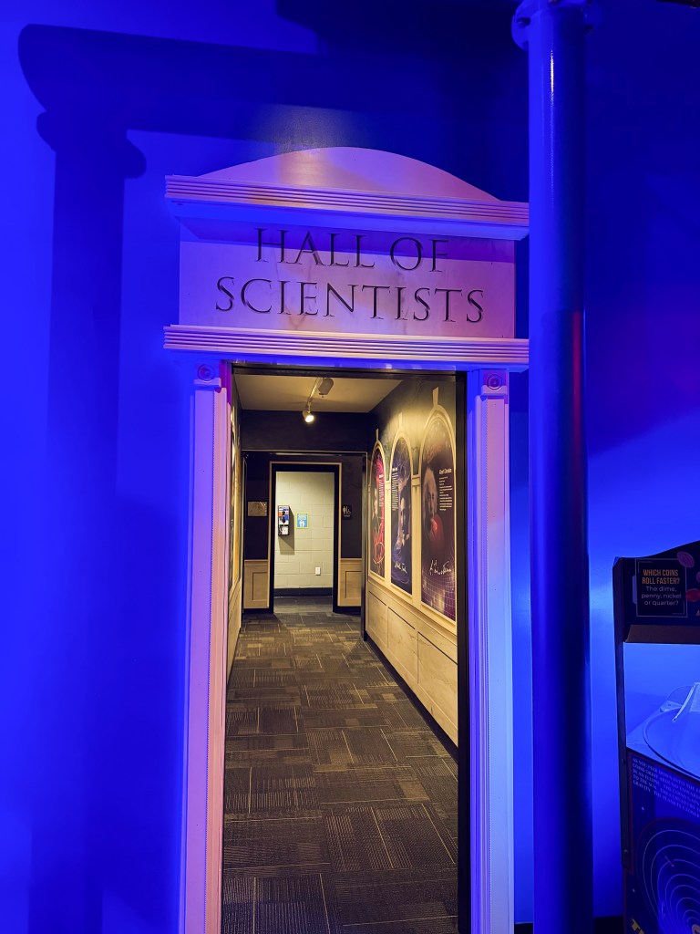 Entrance to a brightly lit hallway titled 'Hall of Scientists' with blue lighting and informational displays along the walls.