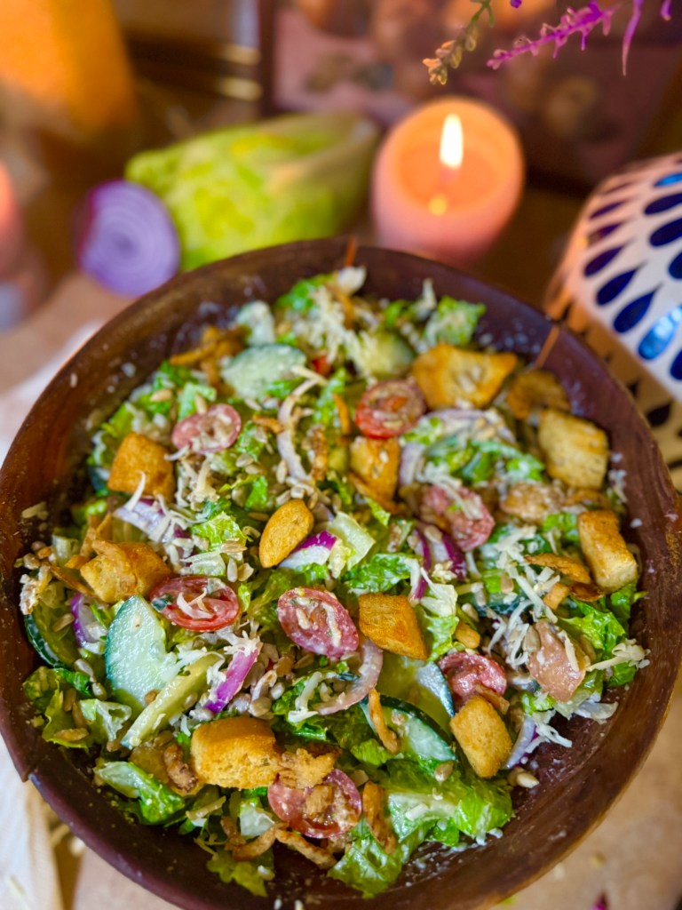 A close-up view of a vibrant salad in a wooden bowl, featuring lettuce, cucumbers, cherry tomatoes, red onion, croutons, and shredded cheese, with candles and decorative items in the background.