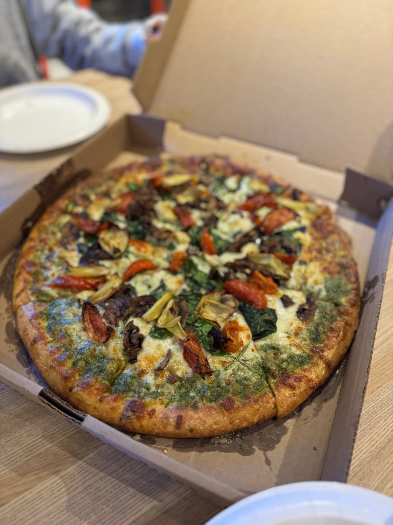 A freshly baked pizza topped with spinach, roasted red peppers, artichokes, and pesto sauce, presented in a pizza box on a wooden table.