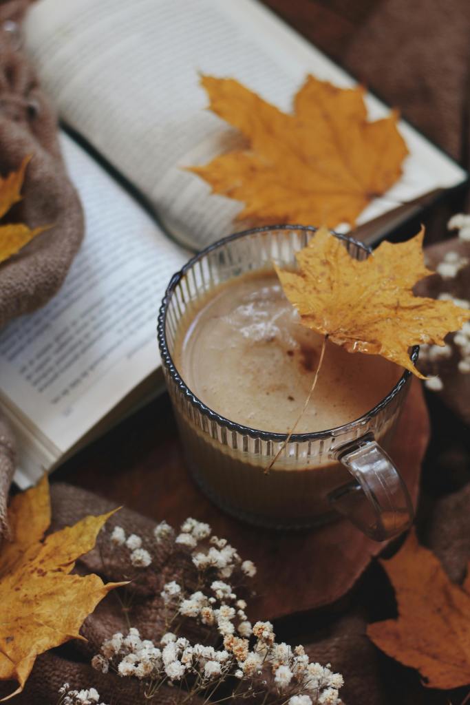 A cozy autumn scene featuring a glass of beverage topped with an orange leaf, surrounded by yellow leaves, a book, and delicate white flowers on a brown background.