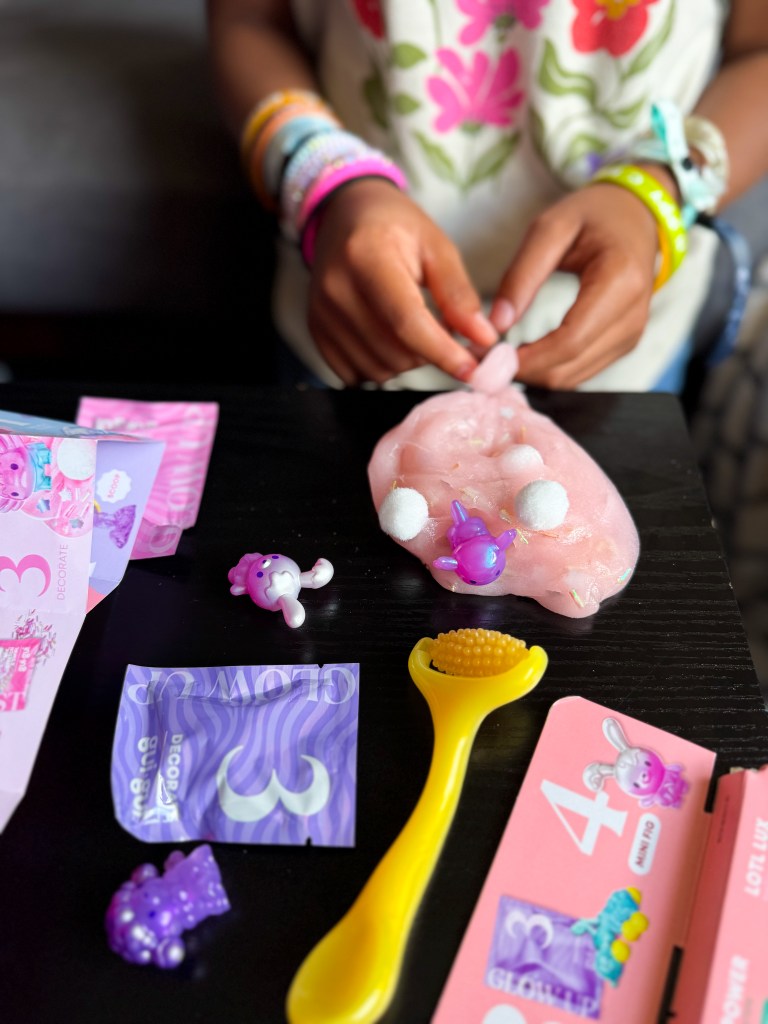 A child playing with pink slime and various colorful plastic toys on a black table. The scene includes small figurines, a yellow tool, and decorative packets featuring playful designs.