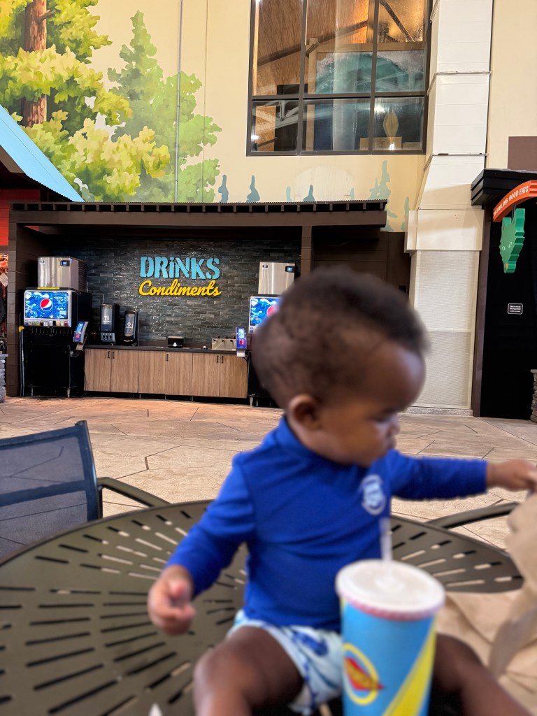 A toddler seated at a table in a resort area, wearing a blue rash guard, with snacks and a colorful drink in front of them. The background features a mural of trees and a counter labeled 'Drinks & Condiments'.
