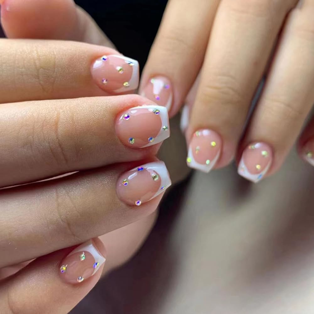Close-up of hands showcasing manicured nails with a nude base and white tips, adorned with colorful rhinestones.