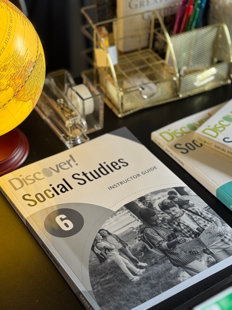 A close-up view of a globe and an open Instructor Guide for Discover! Social Studies Grade 6, with pens and a notepad visible in the background.