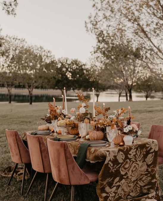 A beautifully styled outdoor Thanksgiving tablescape featuring a floral centerpiece, small pumpkins, and lit candles, set against a serene landscape.