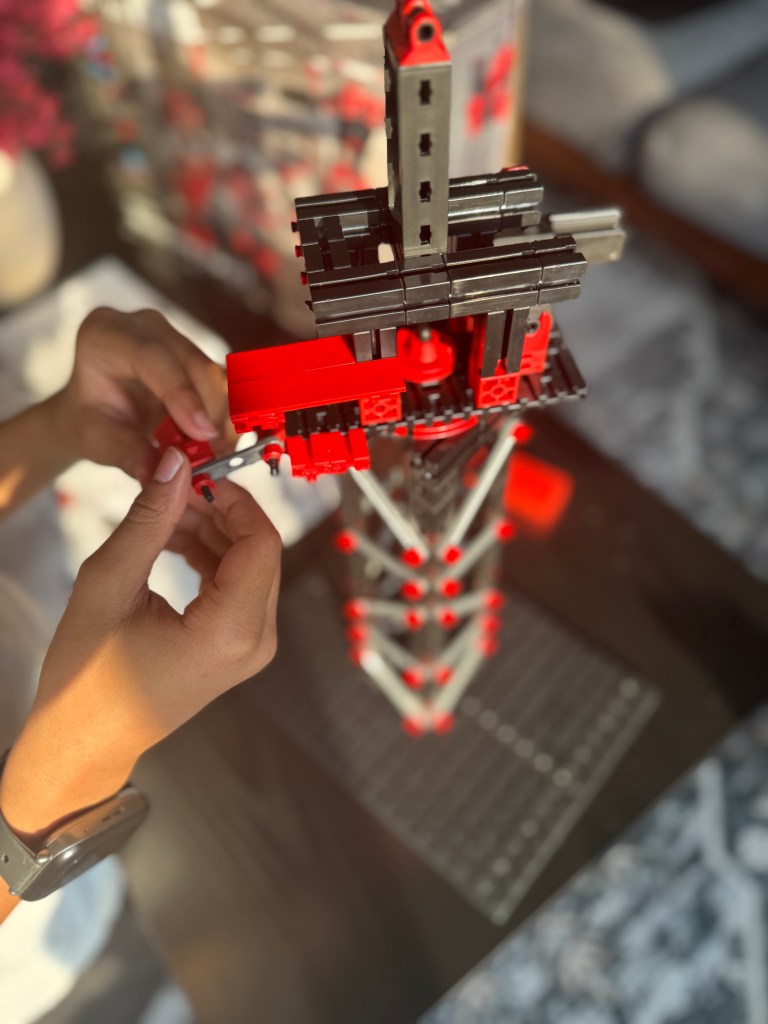 A person assembling a red and black structure using a Fischer Technik STEM kit, with additional pieces visible in the background.