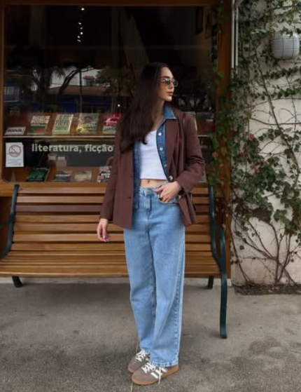 A woman standing outside a bookstore, wearing a brown blazer over a denim shirt and white crop top, paired with light blue wide-leg jeans and sneakers.