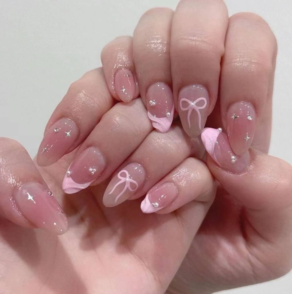 A close-up of hands with stylish nail art featuring a pink French tip design, decorative bows, and small gems on a nude base.