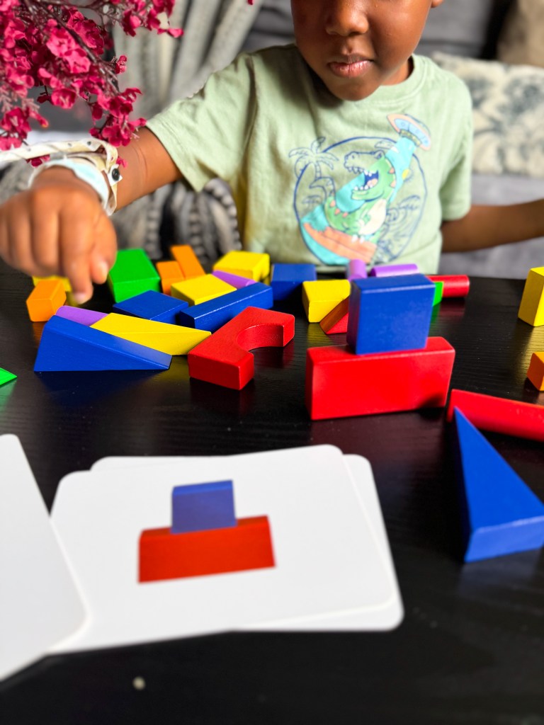 A child plays with colorful geometric building blocks on a table, focusing on arranging shapes next to matching cards.