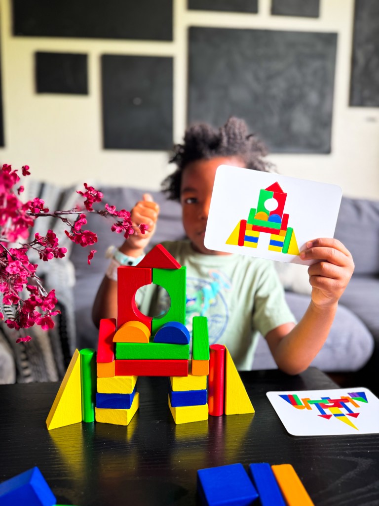 A child holding a card while sitting in front of a colorful block structure made from building blocks. The structure features various shapes and colors, and a pink flower arrangement is on the left side of the image.