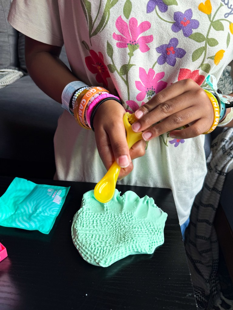 A child playing with mint-green modeling clay and using a yellow spoon to shape it, wearing colorful wristbands, with a blurred background of a cozy indoor setting.