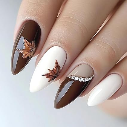 Close-up of elegant almond-shaped nails featuring a mix of dark brown and cream shades. The design includes hand-painted autumn leaves and a delicate rhinestone accent, perfect for a festive Thanksgiving look.