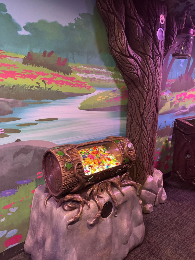 A colorful log-shaped container filled with gummy candies, set against a vibrant forest mural backdrop with flowers and a winding stream.