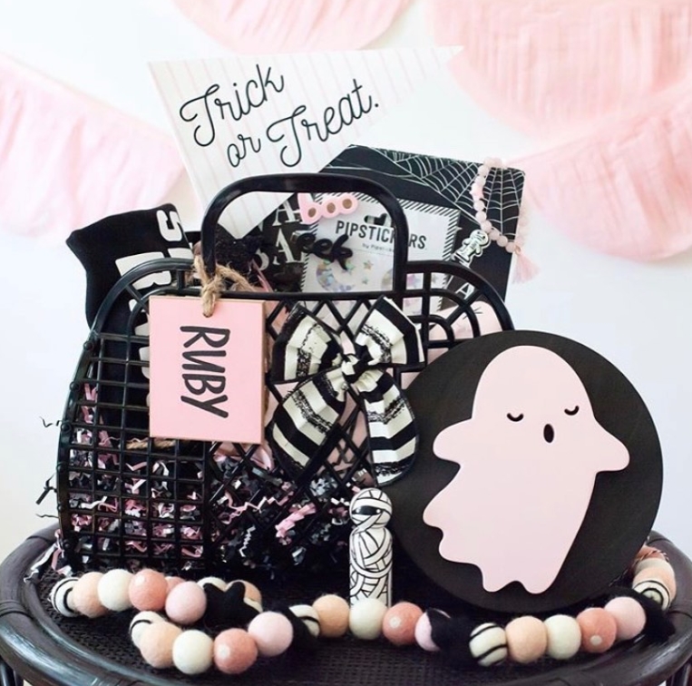 A Halloween-themed basket is filled with treats and decorations, featuring a pink ghost accent, a name tag, and a 'Trick or Treat' banner, surrounded by colorful felt ball garland.