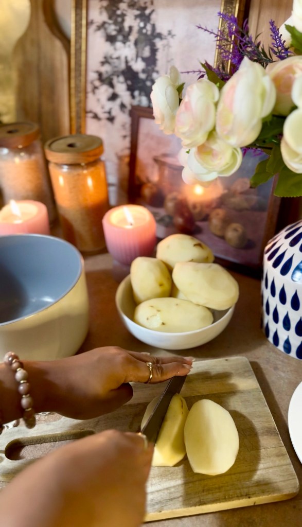 A person slicing potatoes on a wooden cutting board, with a blue bowl and a white bowl filled with peeled potatoes in the background, surrounded by candles and decorative items.