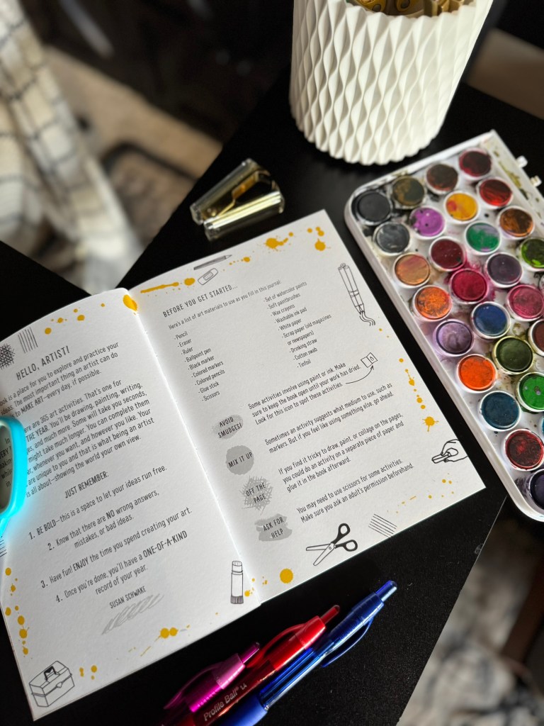 Open art book titled 'My Year of Art' on a table, surrounded by colorful pens and a watercolor palette, with a highlighted page containing prompts for creativity.