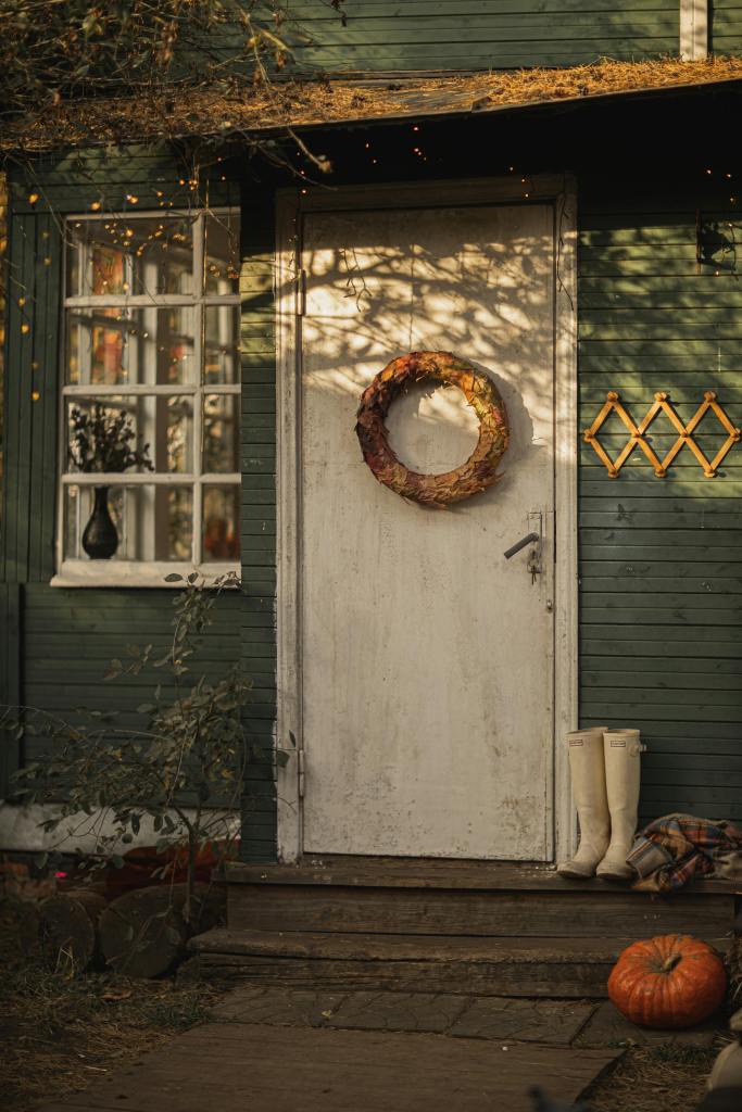 A cozy autumn scene featuring a green wooden door adorned with a wreath made of leaves, a pumpkin by the steps, and rubber boots placed nearby.