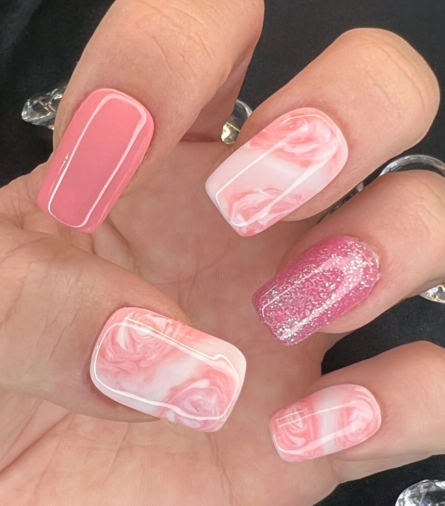 Close-up of a hand with manicured nails featuring a pink and white marble design, a solid pink nail, and a glittery pink accent nail.