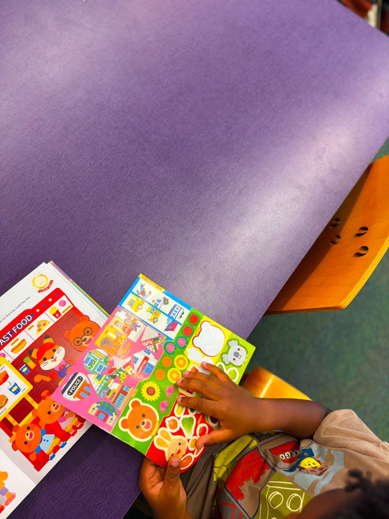 A child is using a colorful sticker book while sitting at a purple table, with a food-themed book visible beside them.
