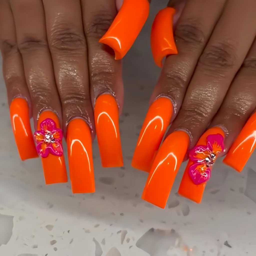 Long neon orange acrylic nails with 3D flower accents and glossy finish