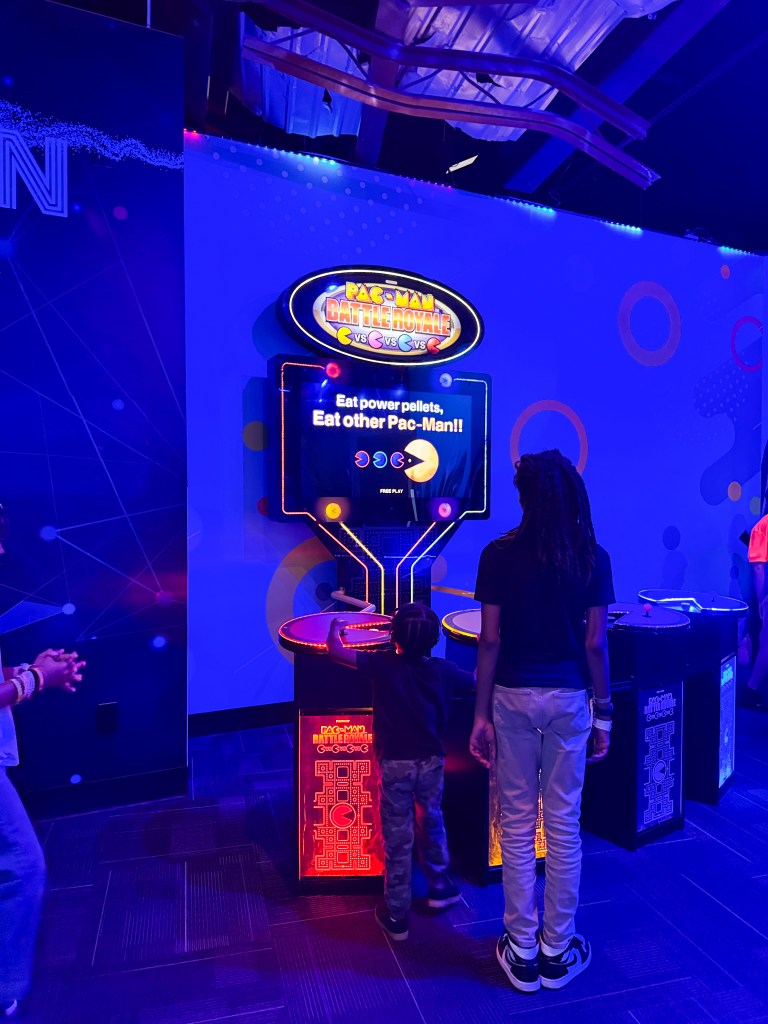 Two children interacting with a 'Pac-Man Battle Royale' arcade game in a brightly lit gaming area.