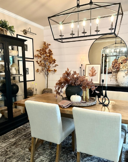A warm and inviting dining room featuring a wooden table set with light-colored chairs. The centerpiece includes a black vase filled with autumn foliage, a white pumpkin, and candles, enhancing the cozy fall atmosphere. A stylish chandelier hangs above, illuminating the space, with framed artwork and a mirror adorning the walls.