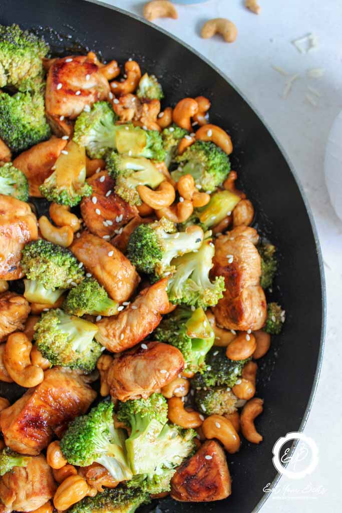 A skillet filled with stir-fried chicken pieces, broccoli, and cashews, topped with sesame seeds.