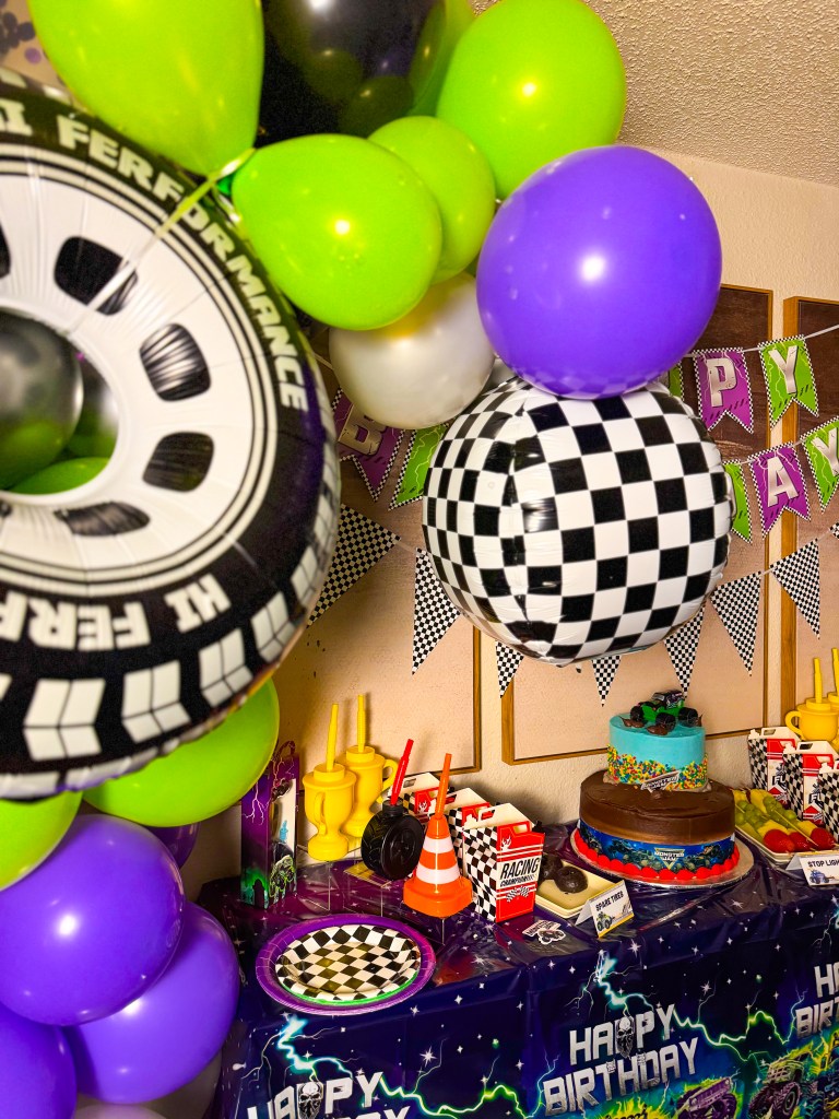 Monster Jam birthday party dessert table with monster truck cake, racing themed decorations, checkered balloons, and themed snacks.