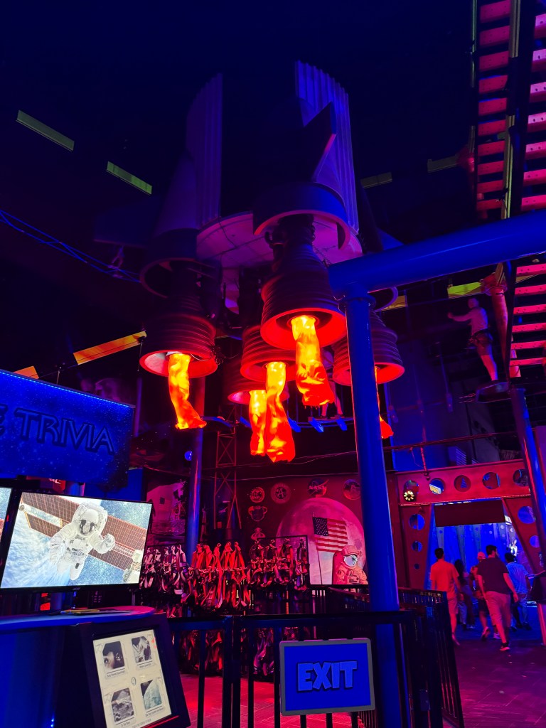 A colorful interior of an attraction featuring rocket engines with fiery lights, screens displaying space-related content, and people walking in the background.
