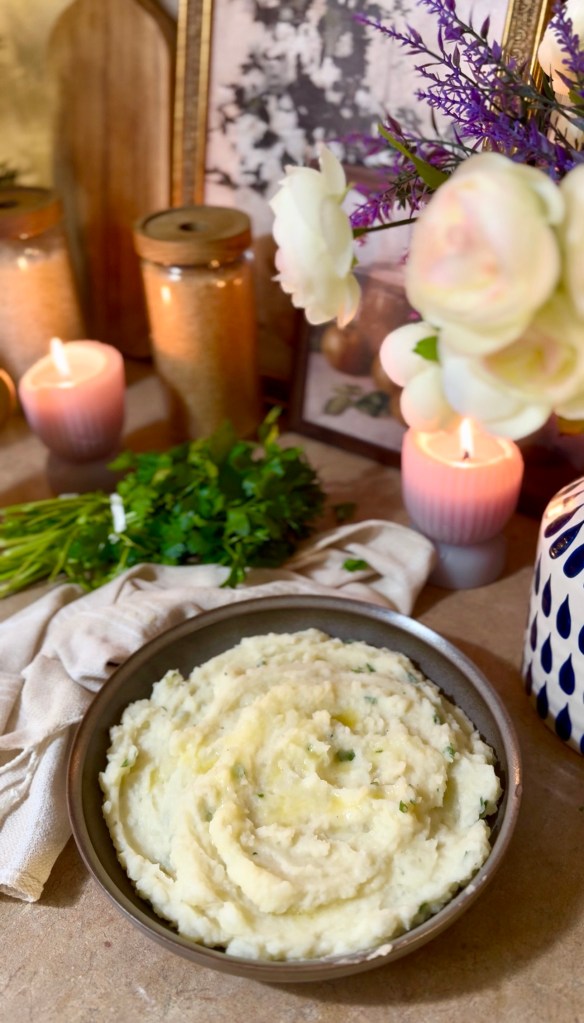 A bowl of creamy mashed potatoes garnished with herbs, surrounded by candles, flowers, and decorative elements.