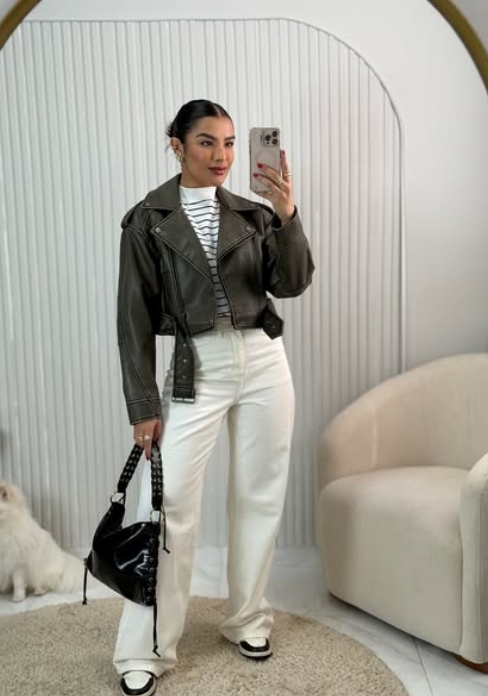 A person posing in front of a mirror wearing a black cropped leather jacket over a striped top and white wide-leg pants, holding a black handbag.
