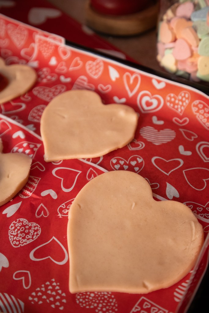 Heart-shaped cookies on red paper with heart patterns, a jar of colorful heart-shaped candies in the background.