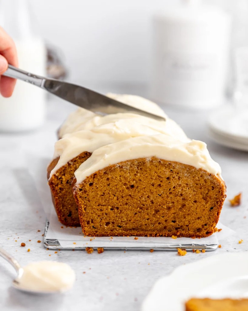 A sliced pumpkin loaf cake topped with cream cheese frosting and a knife spreading frosting on top, placed on a white serving tray with a spoon nearby.