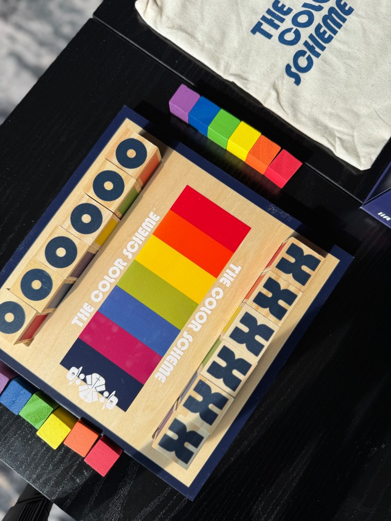 A colorful game board featuring The Color Scheme game with rainbow-colored blocks and code cubes on a black table, accompanied by a canvas storage pouch.
