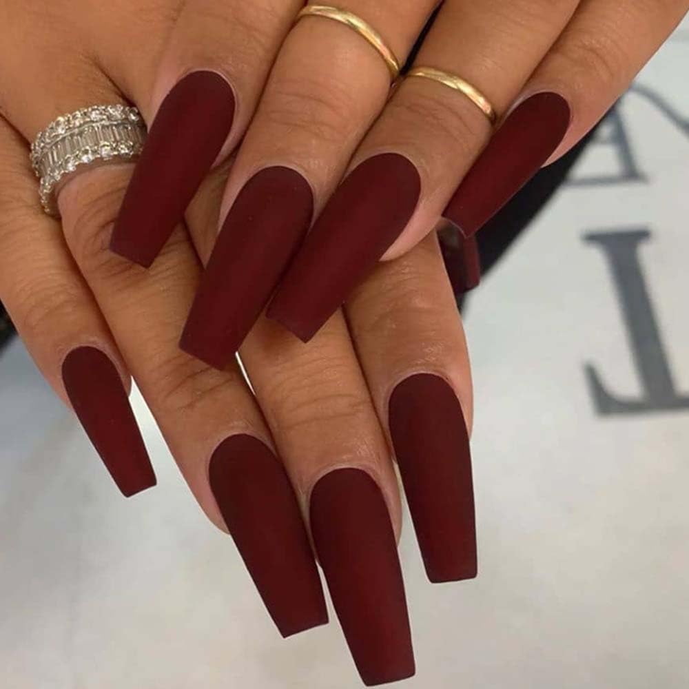 Close-up of hands displaying long, pointed nails painted in a matte burgundy color, adorned with several gold rings.