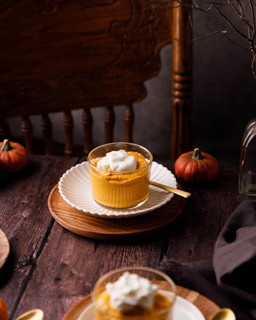 A glass dessert cup filled with pumpkin mousse topped with whipped cream, placed on a white plate, set on a wooden table with small pumpkins in the background.