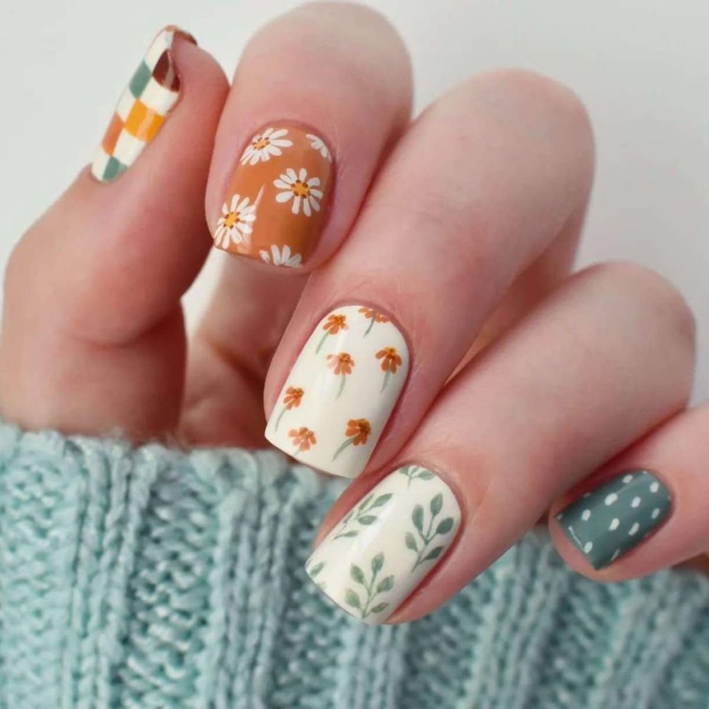 A close-up of a hand showcasing colorful manicured nails with various designs, including floral patterns, checkered print, and pastel colors.