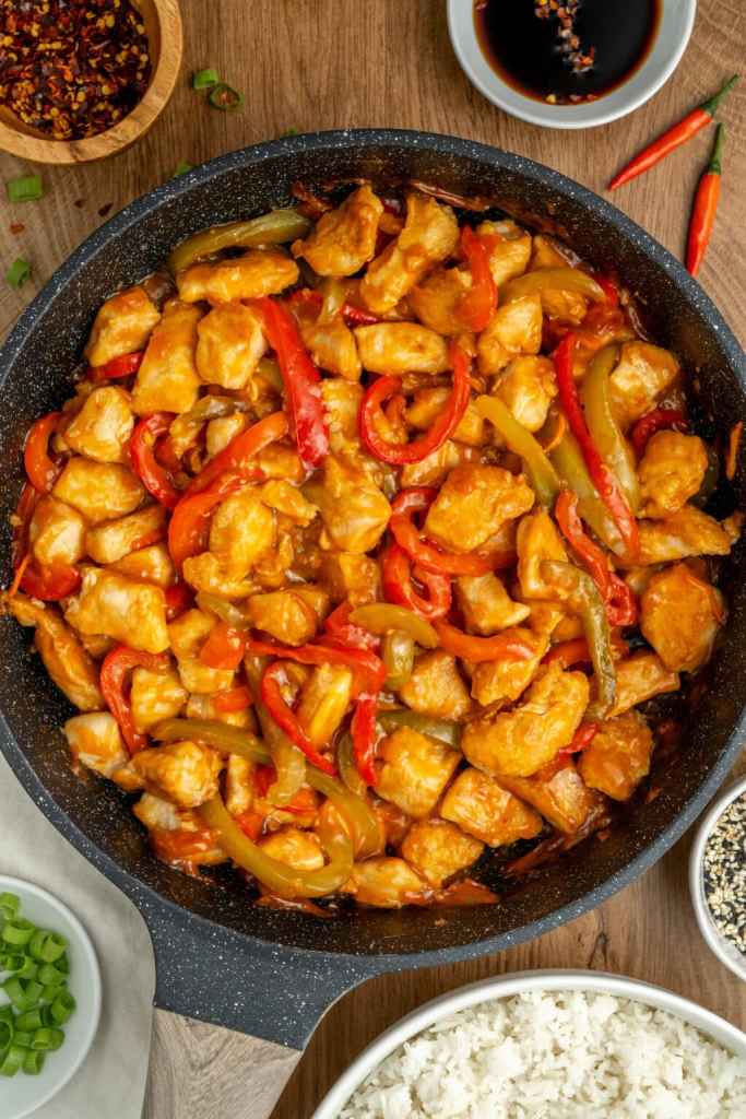 A skillet filled with chicken pieces cooked in a flavorful sauce, combined with sliced red and green bell peppers, garnished with chili and green onions, served alongside bowls of white rice and soy sauce.