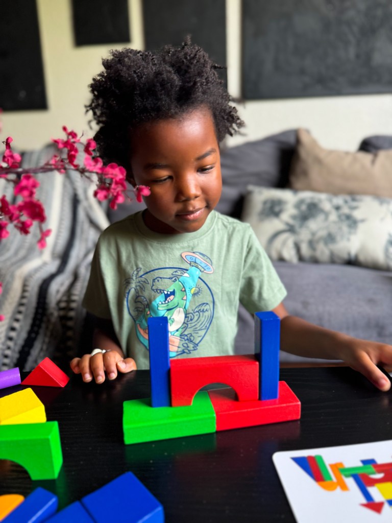A young child with curly hair playfully arranging colorful building blocks on a table, focusing on a red and blue structure they are creating.