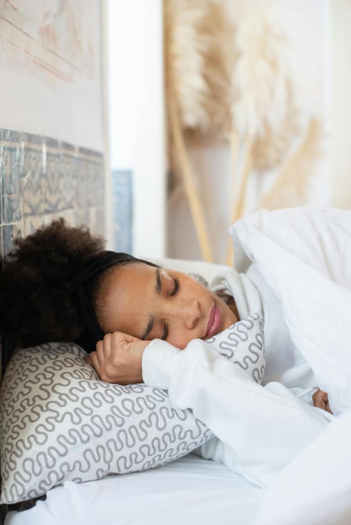 A person with curly hair resting peacefully on a pillow, wearing a cozy sweatshirt, in a softly lit bedroom.